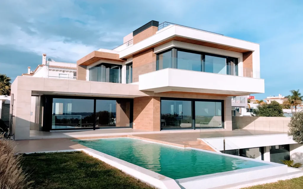 Una casa moderna de dos plantas con grandes ventanales y balcones, que cuenta con una piscina en el patio trasero, rodeada de un césped bien cuidado y palmeras.